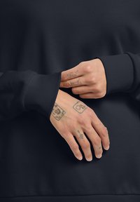 Felpa blu scuro con polsini a costine, che mostra mani con tatuaggi visibili, parzialmente infilate nel dettaglio delle maniche, texture liscia.