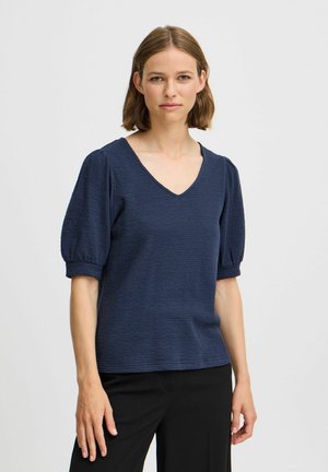 Top in blu navy con texture, scollo a V e maniche corte a palloncino, abbinato a pantaloni neri. Il tessuto sembra leggero e leggermente elasticizzato.