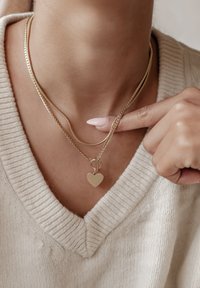 Gelaagde gouden kettingen met een platte hartbedel. De kettingen hebben een gestructureerd ontwerp en worden gedragen over een lichtgekleurde, gebreide trui.