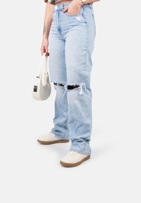 Vaqueros de mezclilla azul claro con un corte relajado, que presentan grandes desgarros deshilachados en las rodillas. Combinados con un bolso blanco y zapatillas beige.