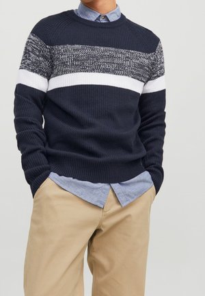 Persona che indossa un maglione a maglia blu navy con strisce bianche e grigie sopra una camicia celeste a bottoni e pantaloni beige con le mani nelle tasche.