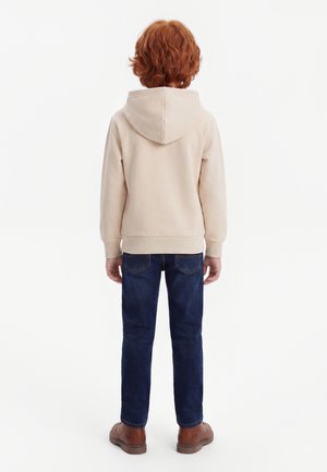 Kind mit lockigem roten Haar, trägt einen hellbeigen Hoodie, dunkelblaue Jeans und braune Schuhe, steht mit dem Rücken zum weißen Hintergrund.