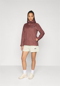 Vrouw die staat met één hand in de zak, gekleed in een bordeauxrode hoodie, beige shorts, witte sokken en beige sneakers tegen een effen achtergrond.