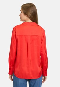 Rote Satinbluse mit Rückenpasse, langen Ärmeln und Knopfmanschetten; zeichnet sich durch eine glatte Textur und einen abgerundeten Saum aus.