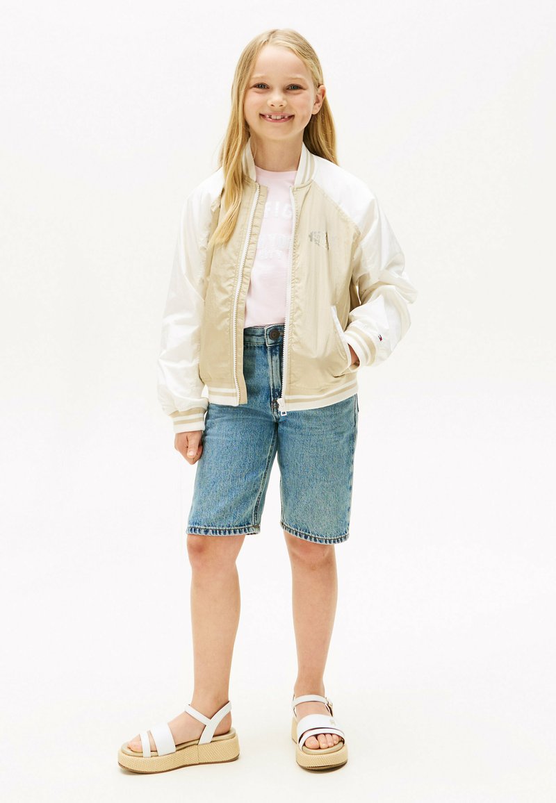 Chaqueta bomber beige y blanca, camiseta rosa, pantalones cortos de mezclilla y sandalias blancas con suelas de plataforma. Tela suave y diseño casual.