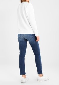 Sudadera blanca con puños y dobladillo acanalados, combinada con jeans ajustados azules y zapatillas blancas. Textura de tela suave y corte casual.