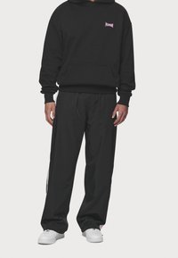 Felpa nera con tasca frontale e logo viola, abbinata a pantaloni neri larghi con strisce laterali e sneakers bianche.