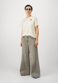 Vaalean beige t-paita, jossa on sininen Nike-logo ja kukon kuvio, yhdistettynä leveälahkeisiin harmaisiin farkkuihin. Asusteina tummat aurinkolasit ja renkaankorvakorut.