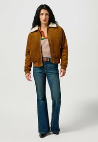 Giacca bomber in velluto a coste marrone con colletto in shearling crema, abbinata a un top a righe in maglia e jeans a zampa d'elefante blu. Stivaletti neri completano il look.
