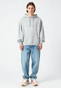 Felpa grigia chiara con tasca frontale, cappuccio con cordini e spalle scese. Abbinata a jeans baggy blu chiaro e sneakers multicolori.