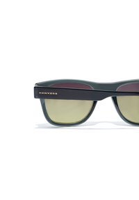 Ochelari de soare din plastic verde cu o ramă groasă, lentile cu un gradient și textul auriu "HAWKERS" pe tija neagră.