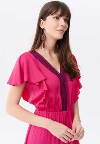 Fuchsia Kleid mit rüschierten kurzen Ärmeln und tiefem V-Ausschnitt, akzentuiert von dunkler Spitzenborte. Trägt große, kunstvoll gestaltete Ohrringe mit Tropfen details.
