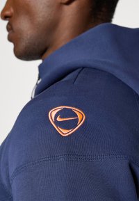 Marineblå hettegenser med teksturert stoff, glidelåslukking og oransje Nike-emblem på venstre skulder. Sømmer er synlige langs mansjetten.