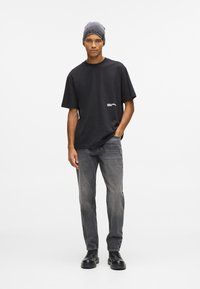 Čierne bavlnené tričko s krátkymi rukávmi a logom na leme, spárované so šedými denimovými džínami a čiernymi šnurovacími čižmami. Nosená sivá čiapka.