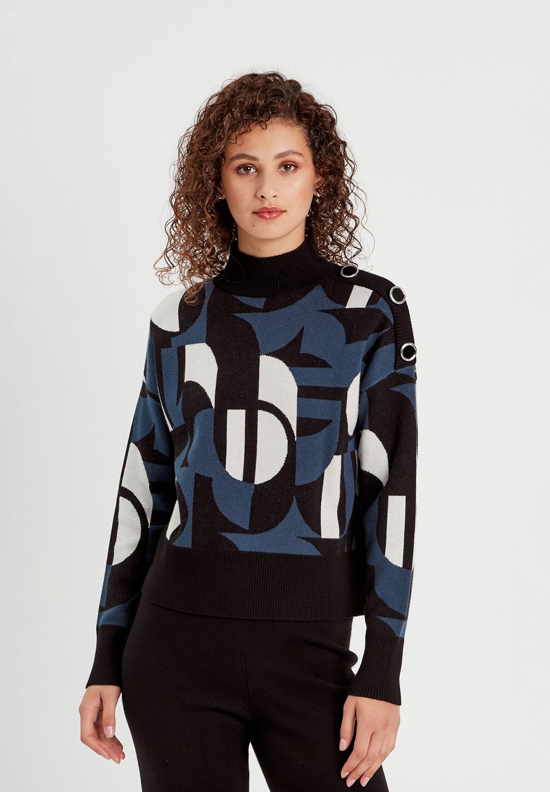 Pull en tricot coloré avec des motifs géométriques en noir, bleu et blanc, munis d'un col montant et de quincaillerie décorative sur les épaules.