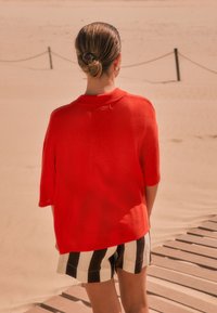 Roter, übergroßer Kurzarm-Pullover mit weicher Textur, getragen über schwarz-weißen gestreiften Shorts. Im Hintergrund sind eine sandige Oberfläche und Holzplanken zu sehen.