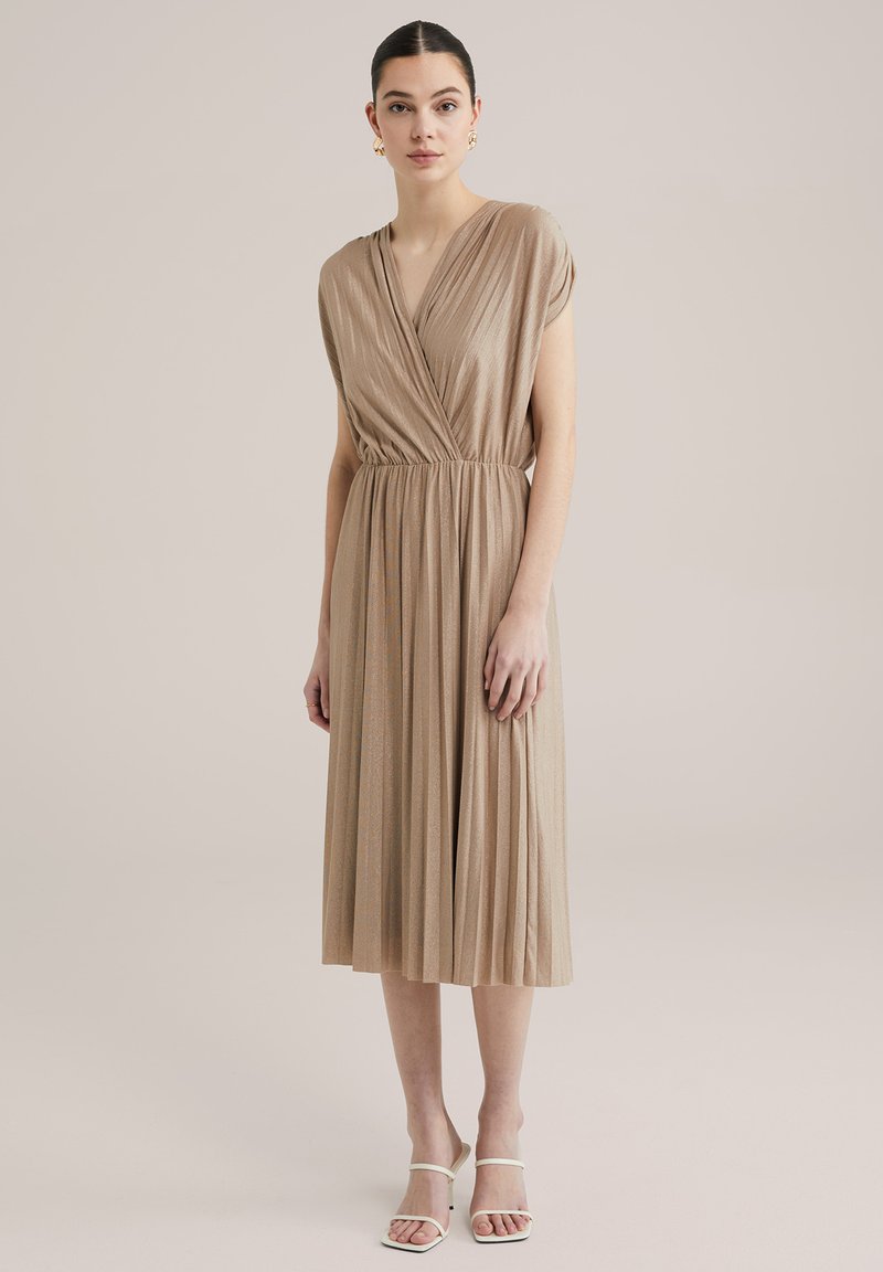 Beige Faltenkleid mit V-Ausschnitt und kurzen Ärmeln, aus einem strukturierten, schimmernden Stoff gefertigt. Getragen mit weißen Riemchensandalen.