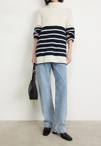 Persona vestida con un suéter a rayas crema y azul marino, jeans de pierna ancha en azul claro, zapatos negros de punta cuadrada y sosteniendo un bolso negro.