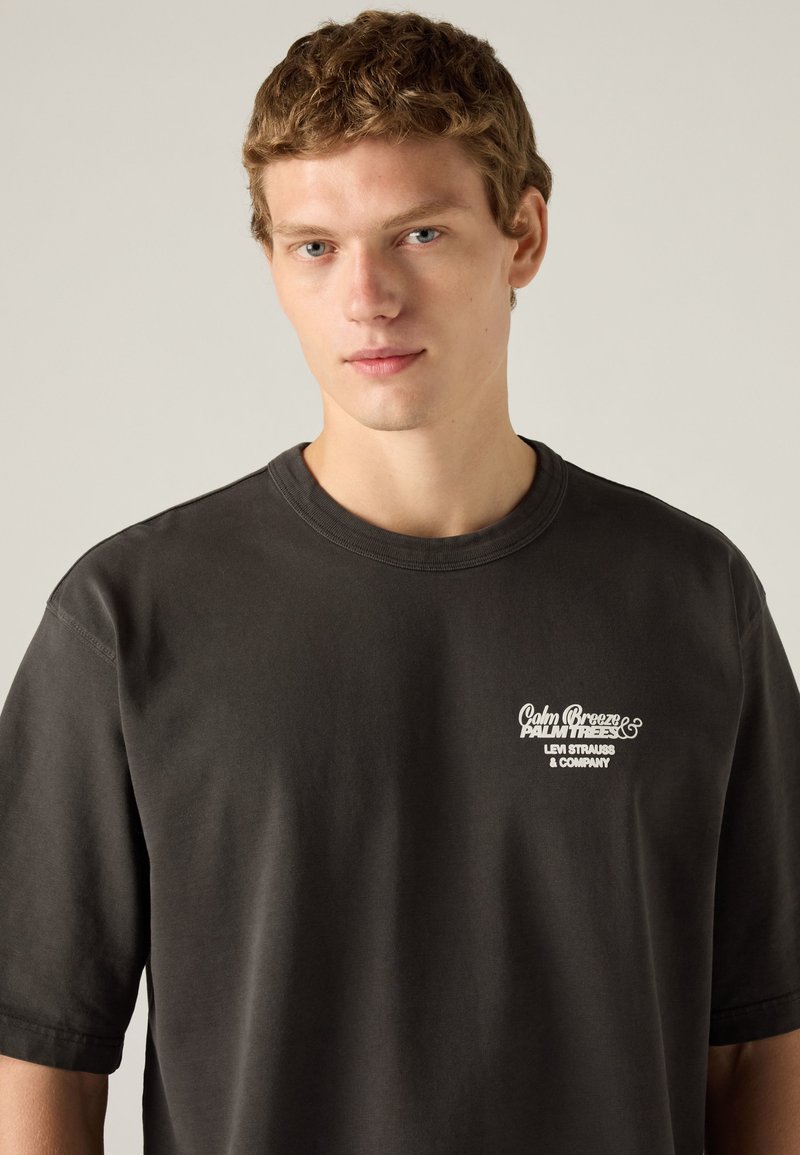 Young man with short curly hair wearing a dark t-shirt with white text reading Calm Breeze Palm Trees Levi Strauss & Company.