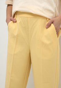 Pantalons larges jaunes en tissu lisse, avec des plis à l'avant, des poches latérales et une taille cintrée avec passants de ceinture.