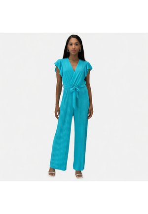 Vrouw staand in een turquoise geplooide jumpsuit met korte volantmouwen en een gestrikte taille, draagt witte open-teen hakken tegen een witte achtergrond.
