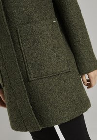 Manteau en laine texturée vert foncé avec grande poche carrée plaquée, manches longues et coutures visibles, porté sur un pantalon noir.