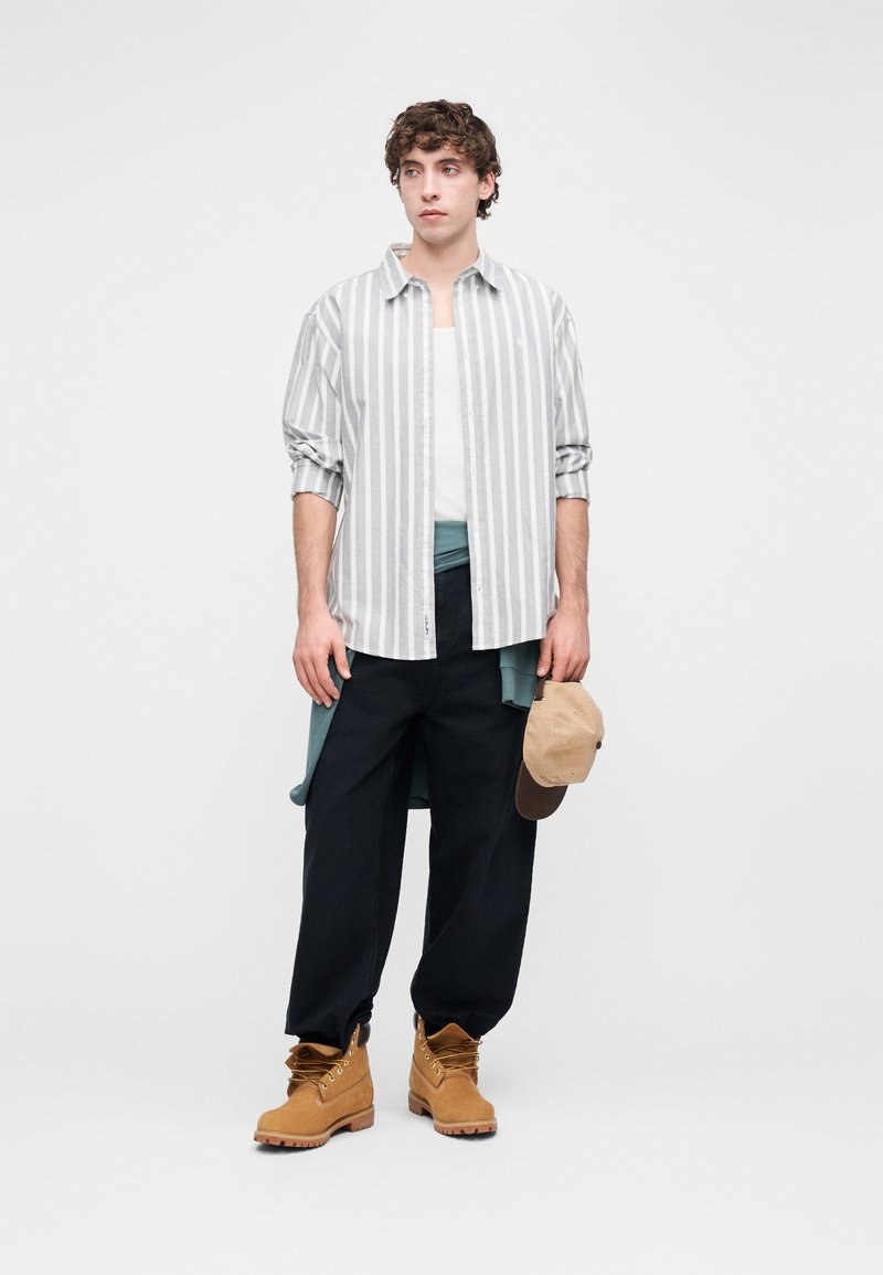 Lysegrå og hvide stribede skjorte, hvid tanktop, mørke bukser, brune støvler, holder en brun kasket. Lagdelt, afslappet design med varierede teksturer.