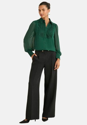 Blusa verde con ricami floreali, maniche lunghe trasparenti; abbinata a pantaloni neri a gamba larghe e tacchi appuntiti neri.