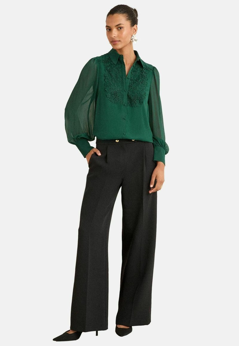 Blusa verde con ricami floreali, maniche lunghe trasparenti; abbinata a pantaloni neri a gamba larghe e tacchi appuntiti neri.