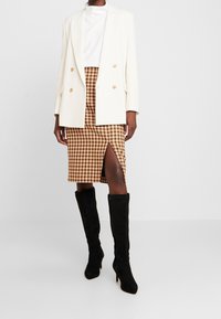 Blazer crema con doppio bottone, top bianco texturizzato, gonna marrone a quadri con spacco laterale, stivali neri in pelle scamosciata al ginocchio.