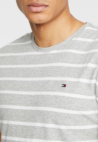 T-shirt gris à rayures présentant des rayures blanches horizontales, un col rond et un petit logo rouge, blanc et bleu sur la poitrine.