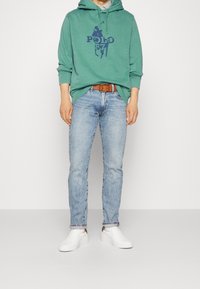 Grüne Kapuzenjacke mit blauem Logo, hellblaue Jeans, brauer Gürtel und weiße Slipper. Lässiger Schnitt, weicher Stoff und entspannendes Design.