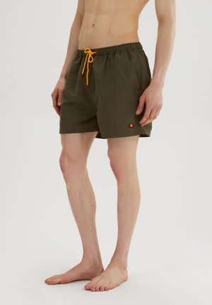 Homme portant un short de bain vert foncé avec un cordon jaune, debout pieds nus devant un arrière-plan clair uni.