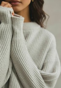 Pull en maille gris clair avec larges manches et texture côtelée, couvrant partiellement les mains. Le tissu présente un aspect doux et confortable.