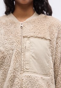 Μπεζ χνουδωτό μπουφάν με υφή εξωτερικό από τεχνητή γούνα, τσέπη από λείο ύφασμα και πλήρες κλείσιμο με φερμουάρ μπροστά.