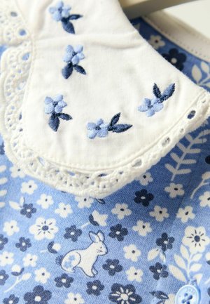 Vaaleansininen kangas, jossa on kukkakuvioita, brodeerattu valkoinen kaulus sinisillä kukilla ja pieni jäniksen kuva.