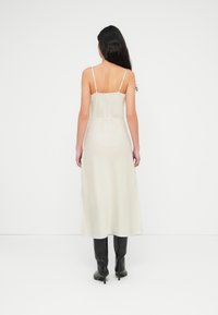 Μακρύ, ανοιχτό κρεμ φόρεμα με λεπτές τιράντες, εφαρμοστή μπλούζα και ρέον φουστάνι. Συνδυάζεται με μαύρες μπότες μέχρι το γόνατο. Λείο, σατινέ ύφασμα.