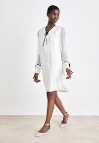 Weißes Kleid mit plissiertem Design, Rüscheldetails und schwarzen Bändern am Ausschnitt. Das Model trägt hellbeige spitze Ballerinas und hat minimalen Schmuck.