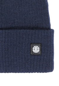 Navy blue knit beanie with a folded cuff and a black label featuring a white logo. Made of soft, textured fabric for warmth.