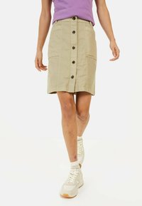 Jupe beige avec fermeture à boutons sur le devant, deux poches et une silhouette droite. Associée à des baskets blanches et une chemise violette.