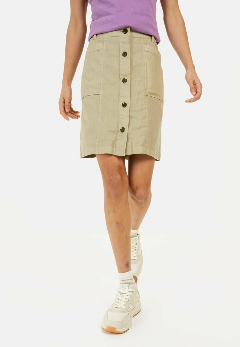 Beige rok met een knoopsluiting aan de voorkant, twee zakken en een rechte silhoette. G gedragen met witte sneakers en een paarse blouse.