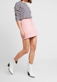 Langärmliges gemustertes Oberteil in Marineblau und Creme, kombiniert mit einem rosa Jeansrock mit vorderen Taschen, ergänzt durch weiße Absatzstiefel.