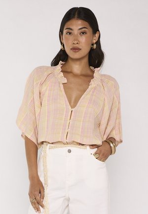 Donna che indossa una camicetta a quadri giallo pastello con scollo arricciato e pantaloni bianchi a vita alta con cintura intrecciata, accessoriata con gioielli dorati.