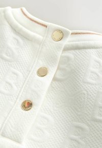 Tejido blanco texturizado con un patrón tejido y letras en relieve. Cuenta con un cuello redondo y tres botones a presión dorados en la tapeta.