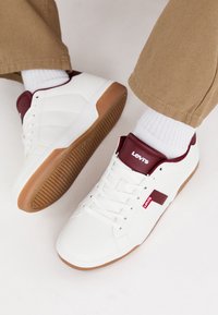 White sneakers with red accents, rubber sole, lace-up design, textured surface; features Levis logo on the tongue and side.