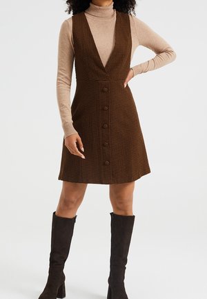 Femme portant une robe sans manches à motifs pied-de-poule marron boutonnée sur le devant, par-dessus un col roulé beige et des bottes noires montant jusqu'aux genoux, se tenant debout avec une main sur la hanche.