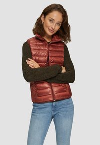 Rote gepolsterte Weste mit hohem Kragen, glänzender Textur, mit Reißverschluss vorne, getragen über einem dunklen, gestrickten Langarmoberteil, kombiniert mit blauen Jeans.