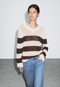 Maglione a righe marroni e crema con scollo a V, vestibilità rilassata, abbinato a jeans a gamba larga blu chiaro. Tessuto strutturato con maniche lunghe.