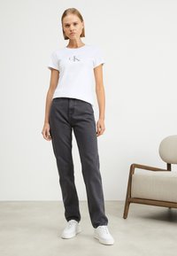 Camiseta de algodón blanca con un logo "ck" gris, combinada con jeans de mezclilla de pierna recta de color gris oscuro y zapatillas blancas. Diseño y ajuste simples.