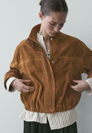 Femme ajustant une veste en daim marron avec des boutons-pression sur une chemise blanche rayée, debout devant un fond clair uni.